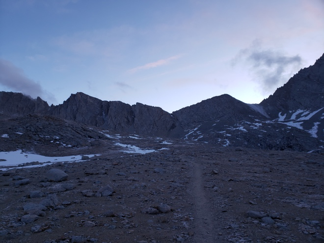 A winding trail leads through lush greenery towards a distant mountain range under a clear blue sky.