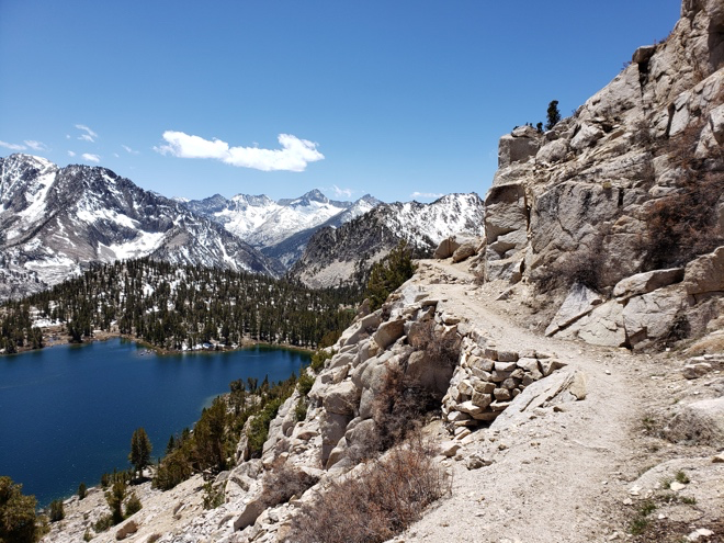 A scenic trail winds up to a serene lake surrounded by majestic mountains in the background.